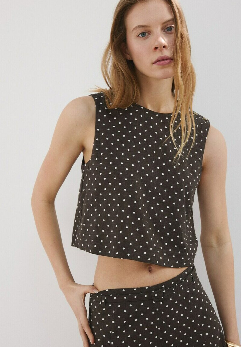 Kobieta z długimi włosami nosi czarny, bezrękawnikowy krótki top i spodnie w białe kropki, pozuje z jedną ręką na biodrze na jednolitym tle.