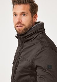 Dunkelbraune Jacke mit hohem Kragen, Reißverschluss vorne und strukturiertem Stoff. Sie hat einen Aufnäher am linken Ärmel und ein weiches Fellfutter.