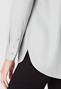 Hellgraues Langarmhemd mit Knopfdetail an den Ärmeln, getragen über schwarzen Hosen, die seitliche Nahtschlitze und einen Teil einer Hand zeigen.