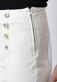 Gros plan sur un pantalon blanc taille haute avec des boutons dorés à l'avant et une fermeture éclair latérale partiellement dézippée, porté avec un haut noir.