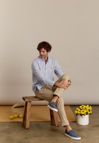 Lichtblauwe overhemdbloes met een klein patroon, beige broek en grijze sneakers. Gestyled zittend op een houten kruk met een bloempot in de buurt.