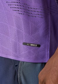 Lila Sportshirt mit Mesh-Struktur, die kleine Löcher und ein geometrisches Muster aufweist. Enthält ein schwarzes ACTIBREEZE-Label am Saum.