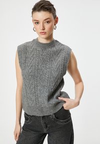 Grauer, kabelstrickartiger ärmelloser Pullover mit hohem geripptem Halsausschnitt; kombiniert mit dunkelgrauen, lässig geschnittenen Jeans mit Seitentaschen.