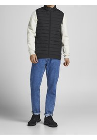 Man med svart vadderad väst över vit långärmad skjorta, blå jeans rullade vid anklarna och svarta sneakers stående mot en neutral bakgrund.