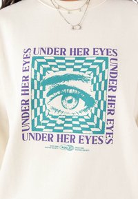 Biely sveter s tyrkysovou grafikou oka v kocovinovom vzore, doplnený textom "UNDER HER EYES" v purpurovej farbe.
