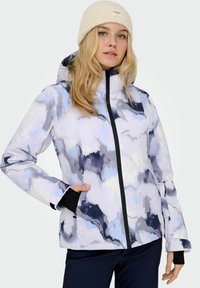 Blond kvinde iført en hvid strikket hue og en vinterjakke med sky-mønster og hætte, sort lynlås og sorte bukser.