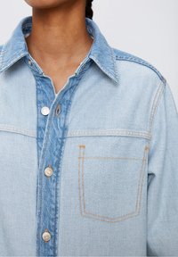 Vaaleansininen farkkupaita, jossa on nappisuljenta edessä, terävä kaulus ja yksi rintatasku. Näkyvät oranssit ompeleet ja metallinapit.