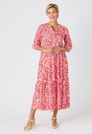 Mujer sonriente de pie con un vestido midi floral rosa y blanco con mangas tres cuartos y sandalias cuña beige sobre un fondo liso.