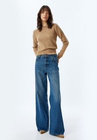 Pull en maille beige et jean bleu à jambes larges. Le pull est à manches longues avec un ourlet côtelé ; le jean est décoloré avec des poches standard et une fermeture à bouton.