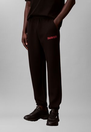 Person iført svarte joggebukser med "Calvin Klein" i rødt på venstre lår, svarte joggesko og svart skjorte, stående med én hånd i lommen.