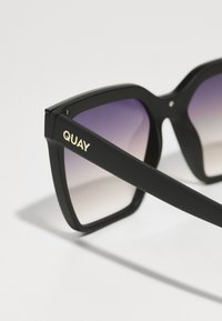 Ochelari de soare negri, pătrați, cu lentile gradient violet. Prezintă logo-ul auriu "QUAY" pe brațul negru. Finisaj lucios cu un design unghiular.