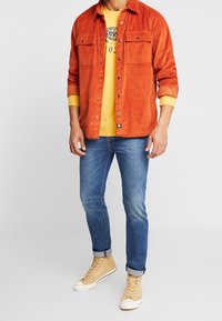 Camisa de pana naranja, camiseta gráfica amarilla debajo, vaqueros azules, zapatillas beige de caña alta. Detalles de botones y bolsillo visibles.