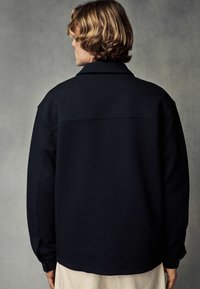 Chemise bleu marine, texturée, avec un col, des manches longues et une coupe droite, dotée de détails de boutons aux poignets.