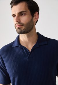 Marineblå ribbet polo med korte ermer og krage med to knapper. Stoffet har en teksturert overflate og en subtil glans.