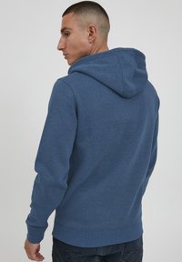 Hættebluse i blåt stof med en tekstureret overflade, ribbede manchetter og kant, afslappet pasform og synlige syninger.