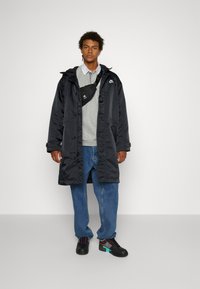 Imperméable noir en nylon, sweat à capuche gris, jean bleu, baskets noires avec des accents turquoise et rose, et sac bandoulière noir. Design simple.