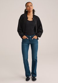 Kvinna med lockigt hår i hästsvans som bär en svart rutig jacka, svart linne, blå jeans och svarta sneakers, står med händerna i fickorna.