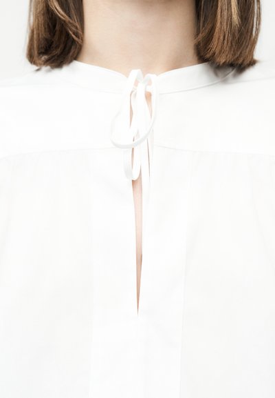 Femme portant un chemisier blanc avec un col noué en boucle, laissant voir une partie de son cou et ses cheveux bruns mi-longs.