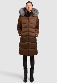 Rjava puhovka z zadrgo spredaj, z oblogo iz krzna na kapuci, vodoravno prešita in stranskimi žepi. Nošeno s črnimi gležnjarji.