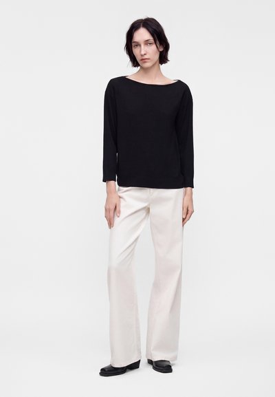 Femme aux cheveux courts et foncés portant un haut noir à manches longues, un pantalon blanc à jambes larges et des chaussures noires, debout devant un fond clair uni.