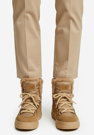 Beige winterlaarzen met bontvoering en dikke veters, gedragen met beige broek, gezien van voren net onder het midden van de kuit.