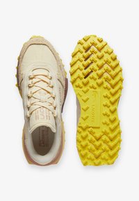 Las zapatillas Lacoste presentan una parte superior beige con texturas de ante y malla, suela de goma amarilla con tacos en forma de zigzag y un detalle de cuero marrón.