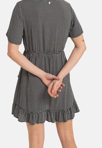 Nainen, joka pitää yllä mustavalkoista ruudullista lyhythihaista mekkoa, jossa on röyhelöhelma, seisoo kädet ristissä selän takana.