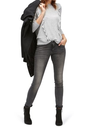 Maglione grigio con maniche a ruffle, abbinato a jeans skinny grigio scuro e stivaletti neri. Un cappotto nero è appoggiato sulla spalla.