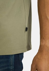 Lys olivenfarvet bomulds t-shirt med lige snit, sideslids og et lille sort mærke ved kanten. Båret med blå denim jeans.