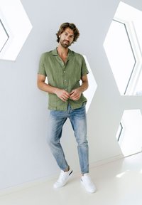 Zelená ľanová košeľa s krátkym rukávom v uvoľnenom strihu, skombinovaná so svetlomodrými džínami a bielymi teniskami, na pozadí moderného geometrického okna.