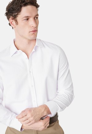 Ung mand justerer manchet på hvid langærmet skjorte med knapper, iført beige bukser og brun bælte, ser mod venstre.