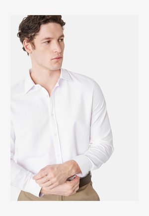 Nuori mies säätää valkoisen pitkähihaisen napillisen paidan kalvosinta, pukeutuneena beigen värisiin housuihin ja ruskeaan vyöhön, katse vasemmalle.