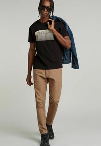 Camiseta negra con un diseño gráfico texturizado, pantalones ajustados beige, chaqueta de mezclilla sobre el hombro y zapatillas negras.