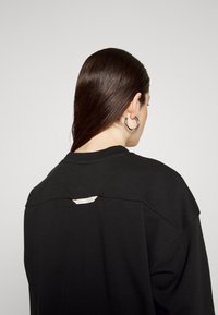 Black sweatshirt with a crew neck, featuring a distinctive tag detail on the back and soft, textured fabric.