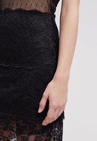 Černé šaty s průhledným korzetem a tečkovanou látkou, zhotovené s jemnými krajkovými detaily na sukně, které zvýrazňují květinové vzory a textury.