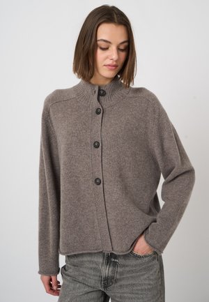 Woman wearing a loose, brown buttoned cardigan with a high collar and faded gray jeans, standing with one hand in pocket.