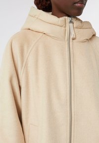 Giacca a cappuccio beige chiaro con una finitura morbida e testurizzata. Presenta una chiusura con zip e un colletto imbottito, con tasche laterali e una vestibilità comoda.