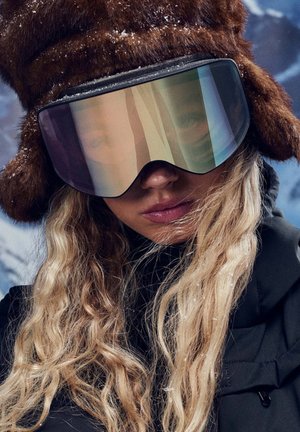 Brūna kažokādu cepure ar ausu aizsegām, kopā ar pārmēru lielām spoguļainām kalnu slēpošanas brillēm. Redzama melna jaka zemā, ar mīkli viļņainiem, gaišiem matiem.