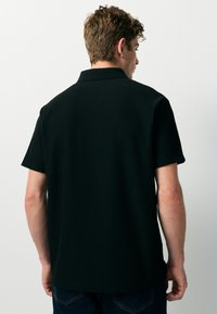 Schwarzes Poloshirt mit kurzen Ärmeln, gerippter Struktur und Kragenansatz, von hinten gezeigt. Minimalistisches Design, ohne sichtbare Logos.