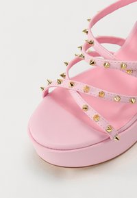 Rosa høyhælte sandal med kryssede stropper og gullmetalliske nagler, avrundet tå og plattformsåle. Jevn tekstur gjennom hele sandalen.