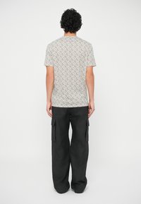 Hombre con pelo rizado vistiendo una camisa de manga corta con estampado y pantalones cargo negros holgados, de pie con la espalda hacia la cámara.