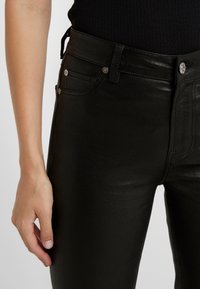 Zbliżenie na osobę w dopasowanych czarnych skórzanych spodniach i czarnej bluzce, z lewą ręką swobodnie opadającą wzdłuż ciała.