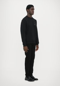 Sudadera negra de tejido texturizado, con cuello redondo y sutiles acentos de logotipo multicolor en el pecho, combinada con pantalones y zapatillas negras.