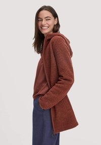 Rote Fleecejacke mit Kapuze, Frontreißverschluss und Seitentaschen; kombiniert mit einem kastanienbraunen Oberteil und blauen Hosen. Glatte Textur, lässiges Design.