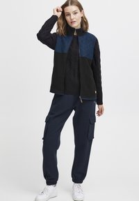 Veste en polaire marine avec un panneau supérieur matelassé, associée à un pantalon cargo marine avec poches. Le modèle porte des baskets blanches.