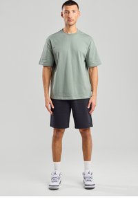 Green cotton t-shirt with a relaxed fit, paired with black shorts. Model wears white athletic shoes with grey accents. Simple design, no patterns.