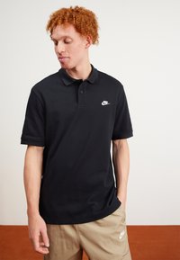 Fekete pólóing, amely pamutból készült, klasszikus gallérral, rövid ujjakkal és fehér Nike logóval a bal mellrészen.