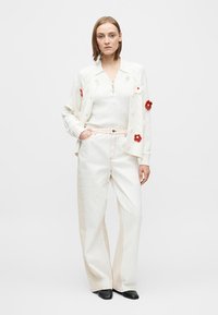 Bijeli pleteni kardigan s crvenim cvjetnim ukrasima, bijeli rebrasti top i svijetle široke hlače. Crne cipele upotpunjuju odjevnu kombinaciju.