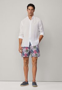 Bijela lanena košulja na dugme, šareni kratke hlače s tropskim uzorkom i plave brodske cipele s smešnim vezicama. Opušteni ljetni outfit, vidljivi detalji.