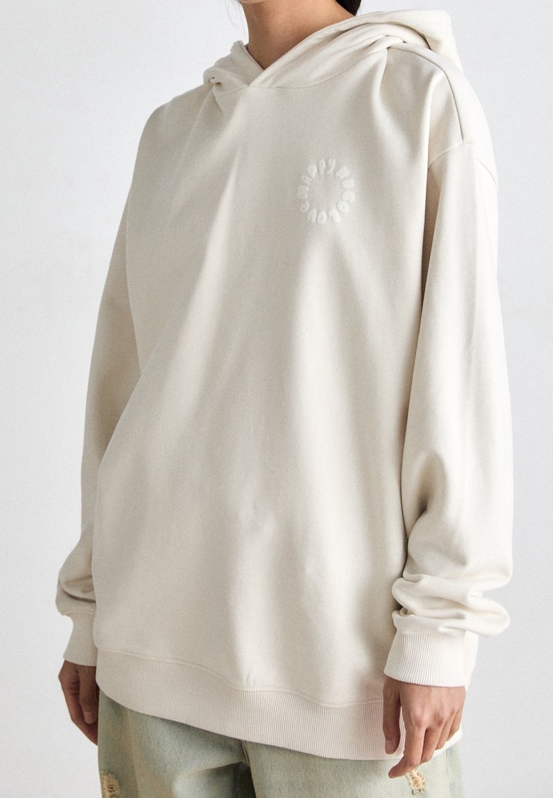Personne portant un sweat à capuche écru avec un logo circulaire subtil sur la poitrine et un jean clair usé, debout devant un fond uni.